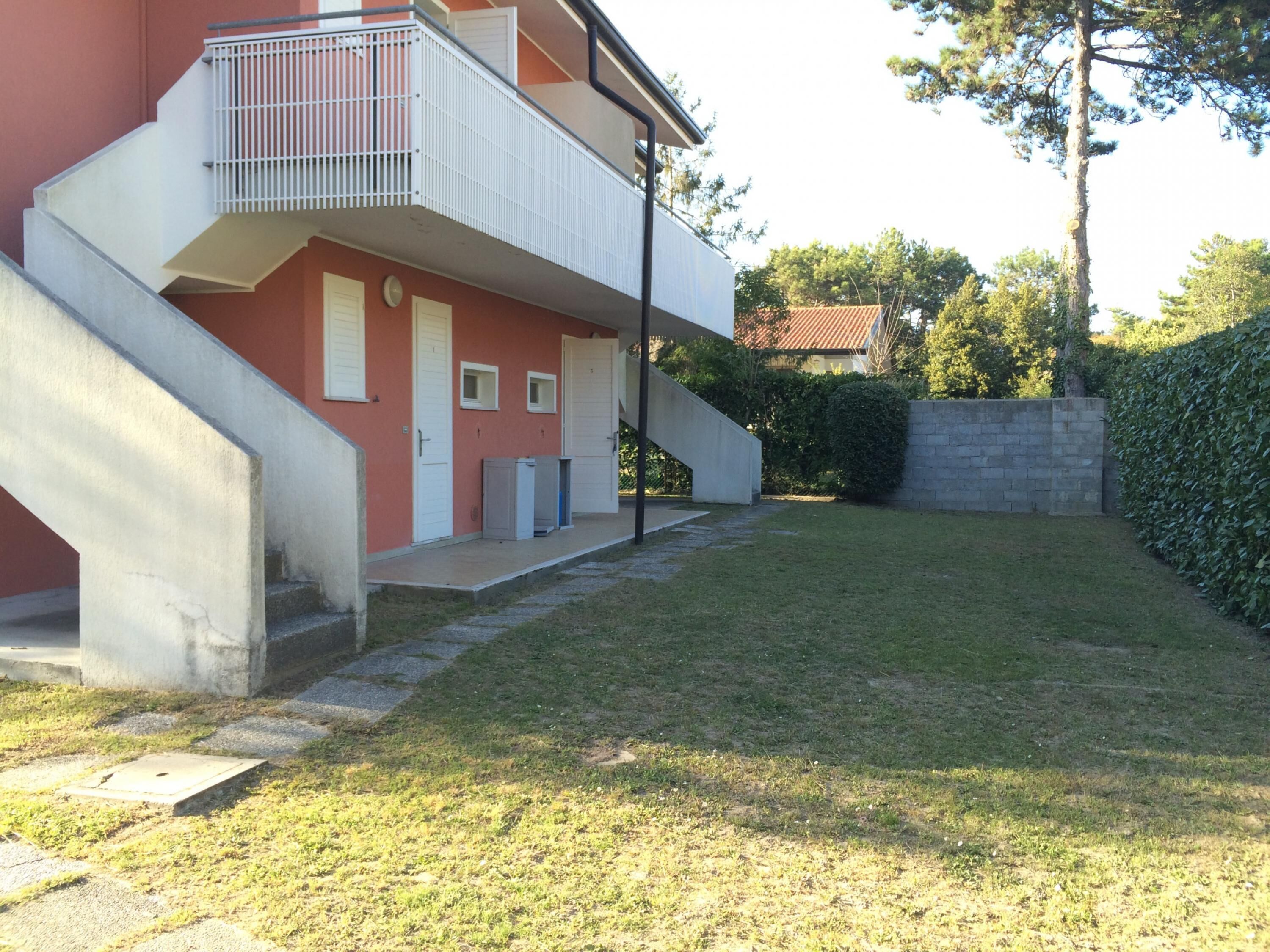 Un bâtiment à deux étages avec une façade rouge, un balcon doté de rampes en métal et un petit jardin agrémenté d’une haie et d’arbres.