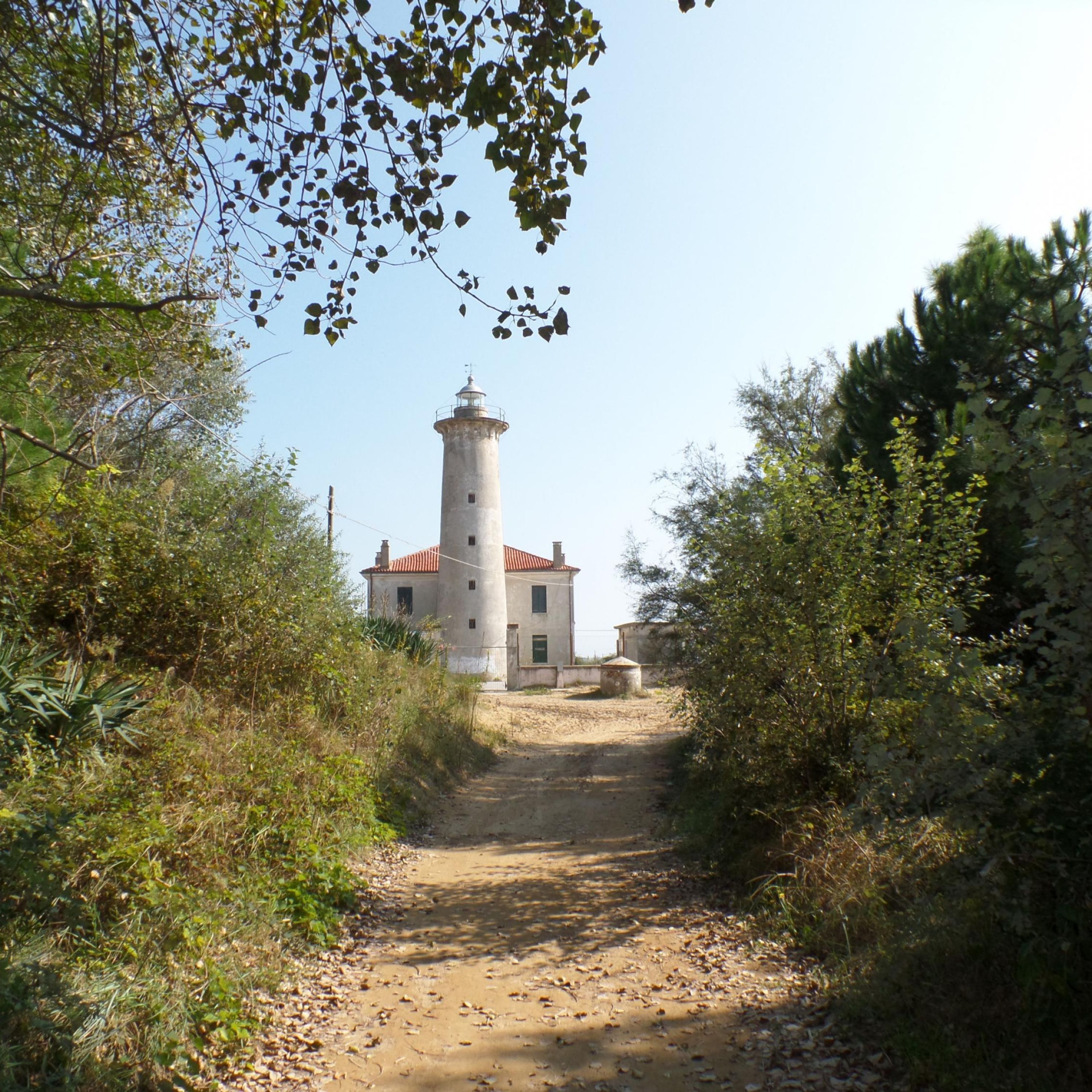 A dirt path leading to a lighthouse with a red roof, surrounded by greenery and trees.
