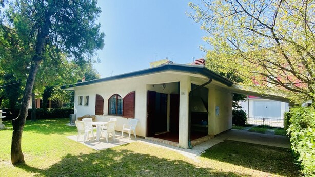A small, single-story house with a covered patio, a small garden, and a white fence. - Rose in Bibione With 3 Bedrooms and 1 Bathrooms (San Michele al Tagliamento)