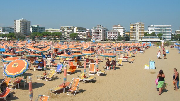A beach with numerous sun umbrellas and beach chairs, a sandy beach, and buildings in the background. - Diana 5 in Bibione With 1 Bedrooms and 1 Bathrooms (San Michele al Tagliamento)