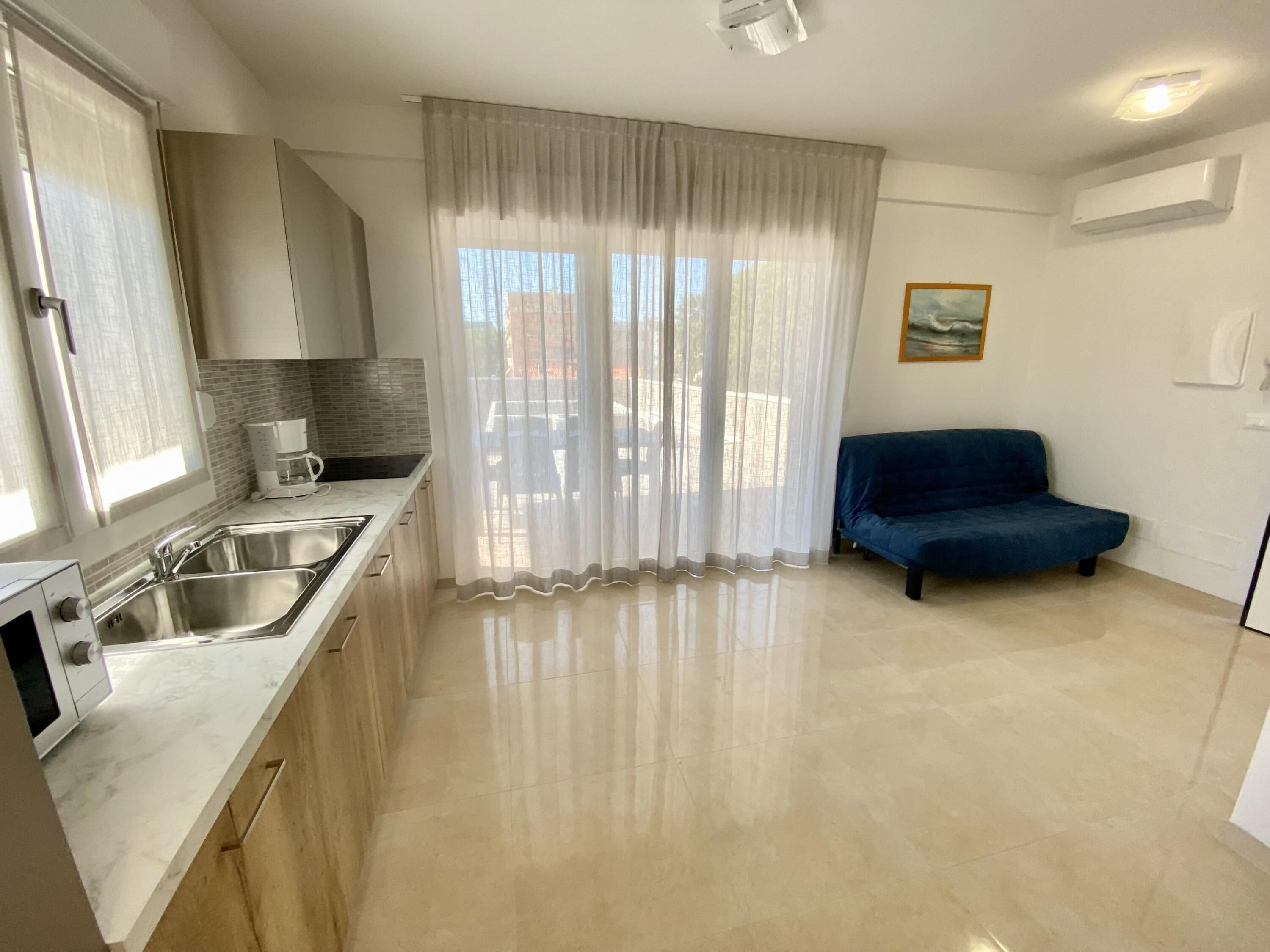 Una cocina moderna con electrodomésticos de acero inoxidable, un sofá azul y un amplio ventanal con cortinas traslúcidas.