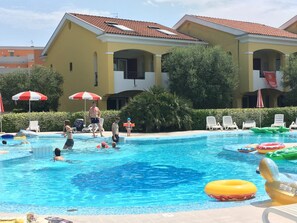 A swimming pool area with lounge chairs and umbrellas, surrounded by a building with a tiled roof. - Borgo E in Bibione With 4 Bedrooms and 2 Bathrooms (San Michele al Tagliamento)