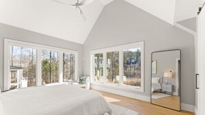 Un'ampia camera da letto con grandi finestre, un letto, un comodino, uno specchio e vista su alberi ed edifici.