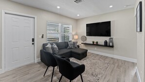 Un salon moderne avec un canapé d’angle, deux fauteuils noirs, un téléviseur à écran plat et une petite étagère ornée d’objets décoratifs.