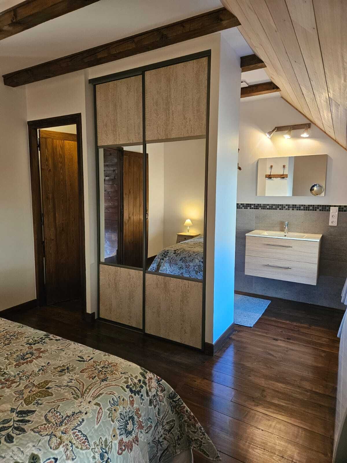 Een slaapkamer met een groot bed, een kledingkast, een spiegel, een nachtkastje met een lamp, en een badkamer met een wastafel en spiegel.