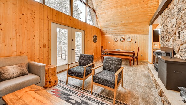 A cozy cabin interior with wooden walls, a sofa, two armchairs, a dining table, and a stone wall.
