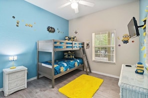 Een stapelbed met een ladder, een klein nachtkastje met een lamp, een raam met jaloezieën en een muur versierd met Minion afbeeldingen.