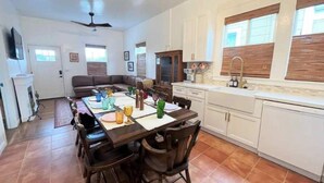 A spacious living room with a large sofa, a dining table set for four, and a kitchen area with white cabinets and a window. - Charming Cottage Retreat (Oakland)