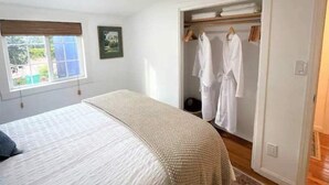 A bedroom with a bed, a window, a framed picture, and a walk-in closet with hanging robes. - Charming Cottage Retreat (Oakland)