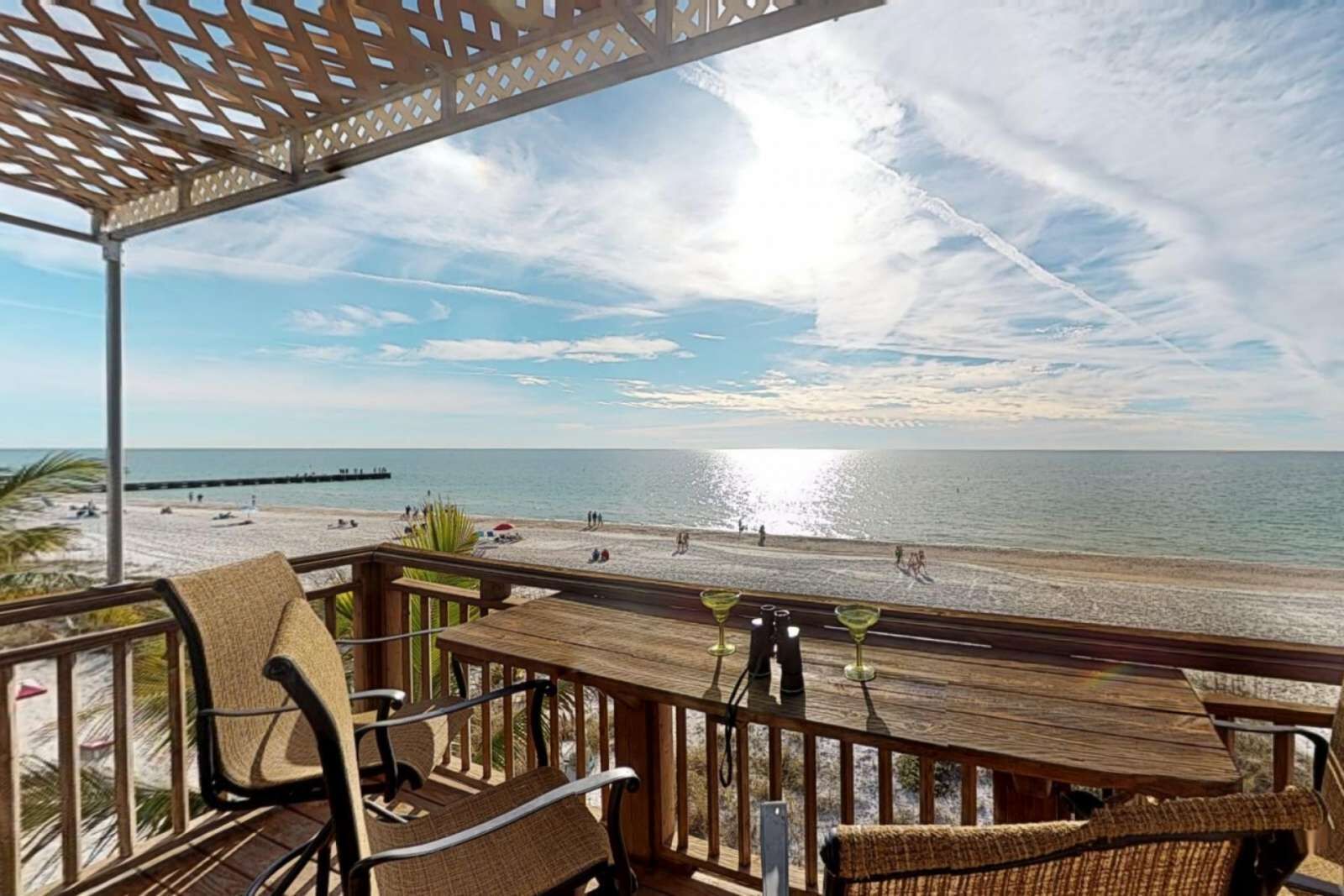 Une terrasse en bois avec des chaises et une table en osier, donnant sur une plage où l’on aperçoit des gens et, au loin, une jetée.