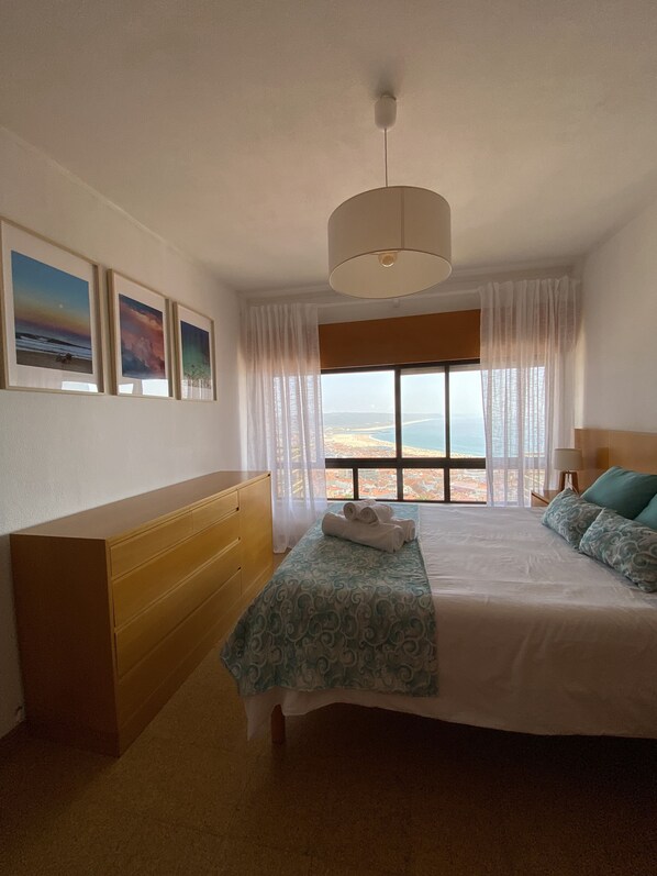 Habitación de hotel con una cama grande, un aparador de madera, un banco y vista al océano.