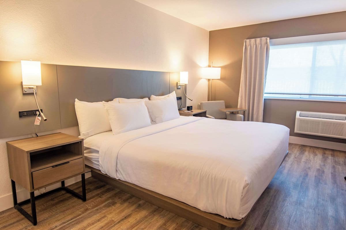 A hotel room with a bed, bedside tables, lamps, a window with curtains, and an air conditioning unit.