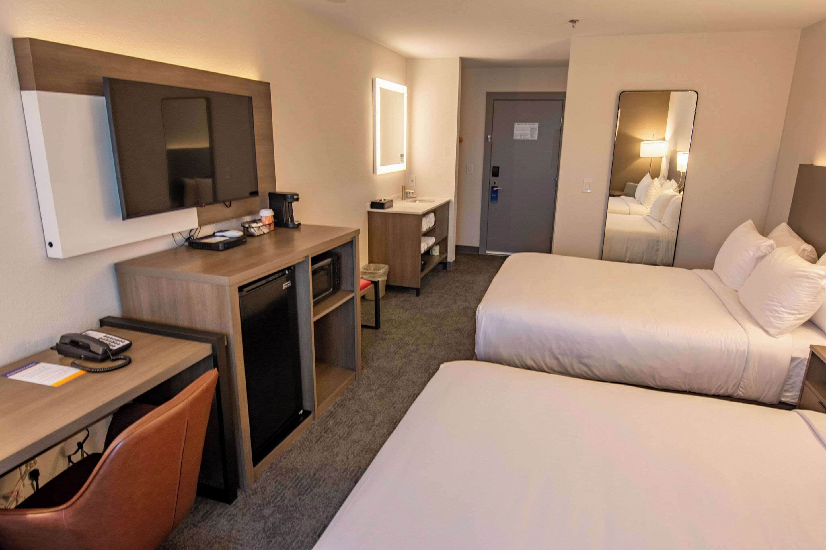 Una habitación de hotel con escritorio, dos camas, televisión, espejo y baño.