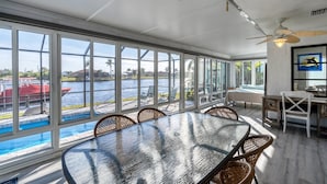 Un solarium con un gran ventanal que da a un cuerpo de agua, una piscina y una zona de comedor con muebles de mimbre.