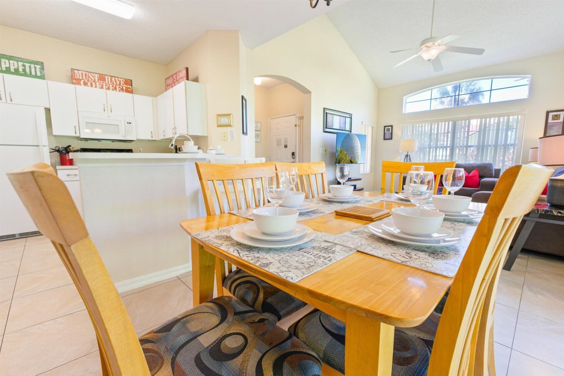 A dining area with a wooden table set for four, surrounded by chairs. There is a kitchen area with white cabinets and a ceiling fan.