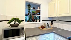 A kitchen with white cabinets, a microwave, a sink, and a window with shelves holding plants and kitchenware. - Charming Home Near Gatwick Airport (Crawley)