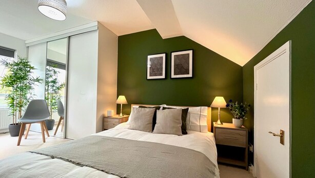 A modern bedroom with a large bed, two bedside tables with lamps, a green accent wall, and a sliding glass door leading to a balcony with plants. - Charming Home Near Gatwick Airport (Crawley)