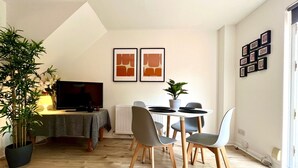A modern living room with a dining area, featuring a TV, a dining table with chairs, a potted plant, and framed artwork on the wall. - Charming Home Near Gatwick Airport (Crawley)