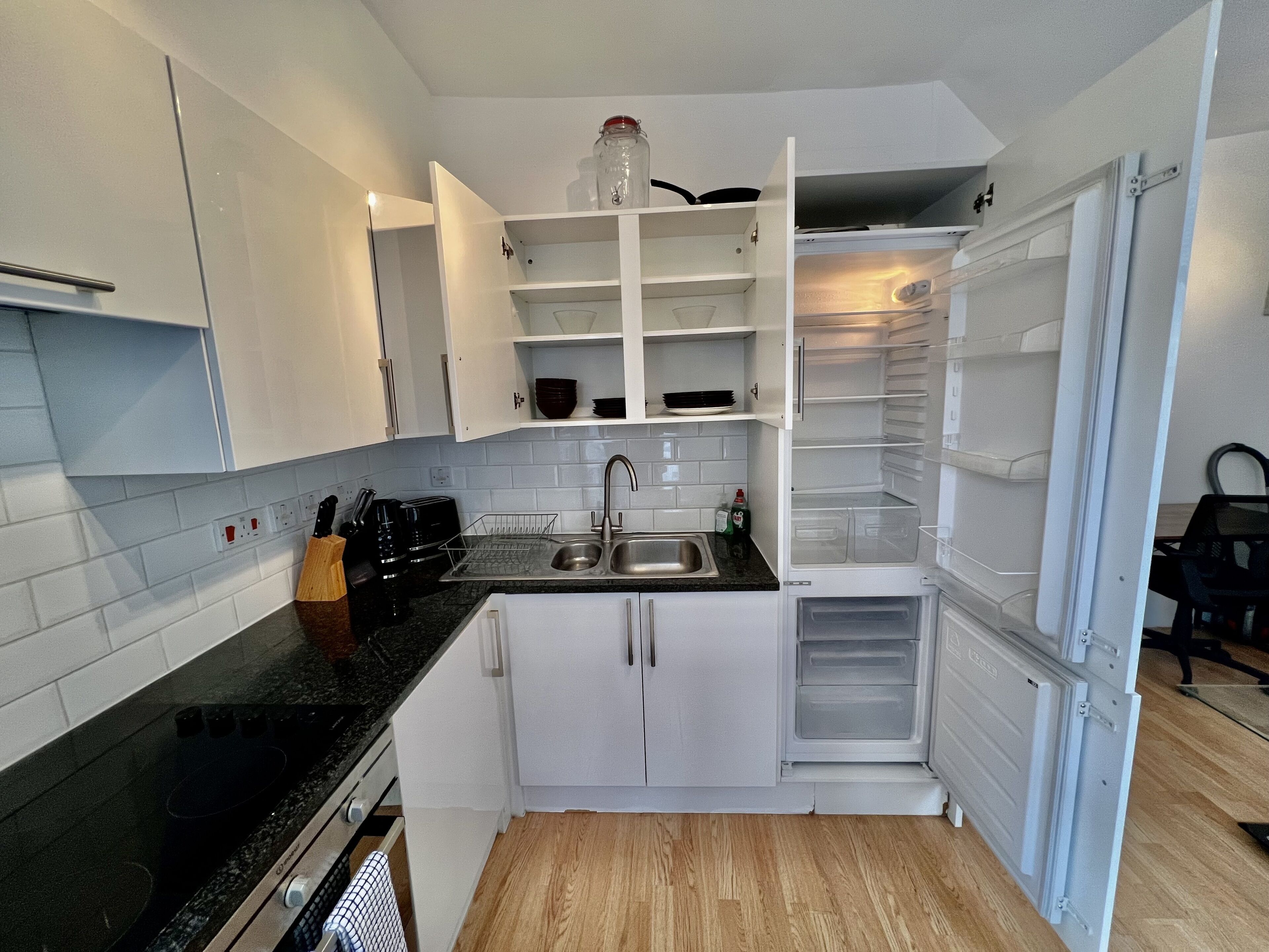 Een kleine keuken met witte kastjes, een zwart aanrecht, een ingebouwde vaatwasser en een koelkast met verschillende compartimenten.