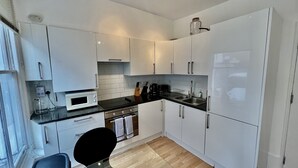 Cocina compacta con armarios blancos, un microondas, un lavamanos y una silla negra.
