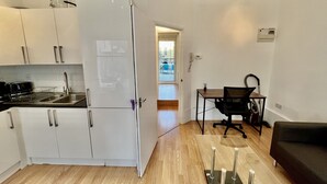 Cocina compacta con gabinetes blancos, encimera negra y refrigerador. Hay una zona de comedor con mesa y sillas, y un sofá negro.