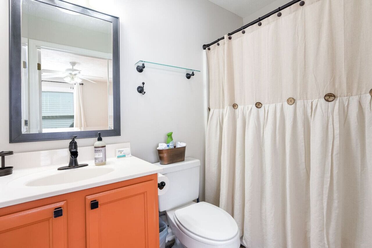 Une salle de bain avec un lavabo blanc, un miroir, un meuble orange, des toilettes et un rideau de douche.