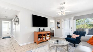 A living room with a flat-screen TV, a coffee table, a sofa, and a chair. There is a door leading to a balcony with a view of trees. - *Family Stay 2 minutes to downtown Sleeps 10 (Auburn)