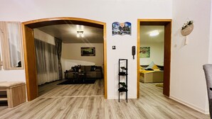 Una sala de estar con piso de madera, un sofá, una mesa de centro y una estantería. Hay una puerta que da a otra habitación con una cama y un cuadro en la pared.