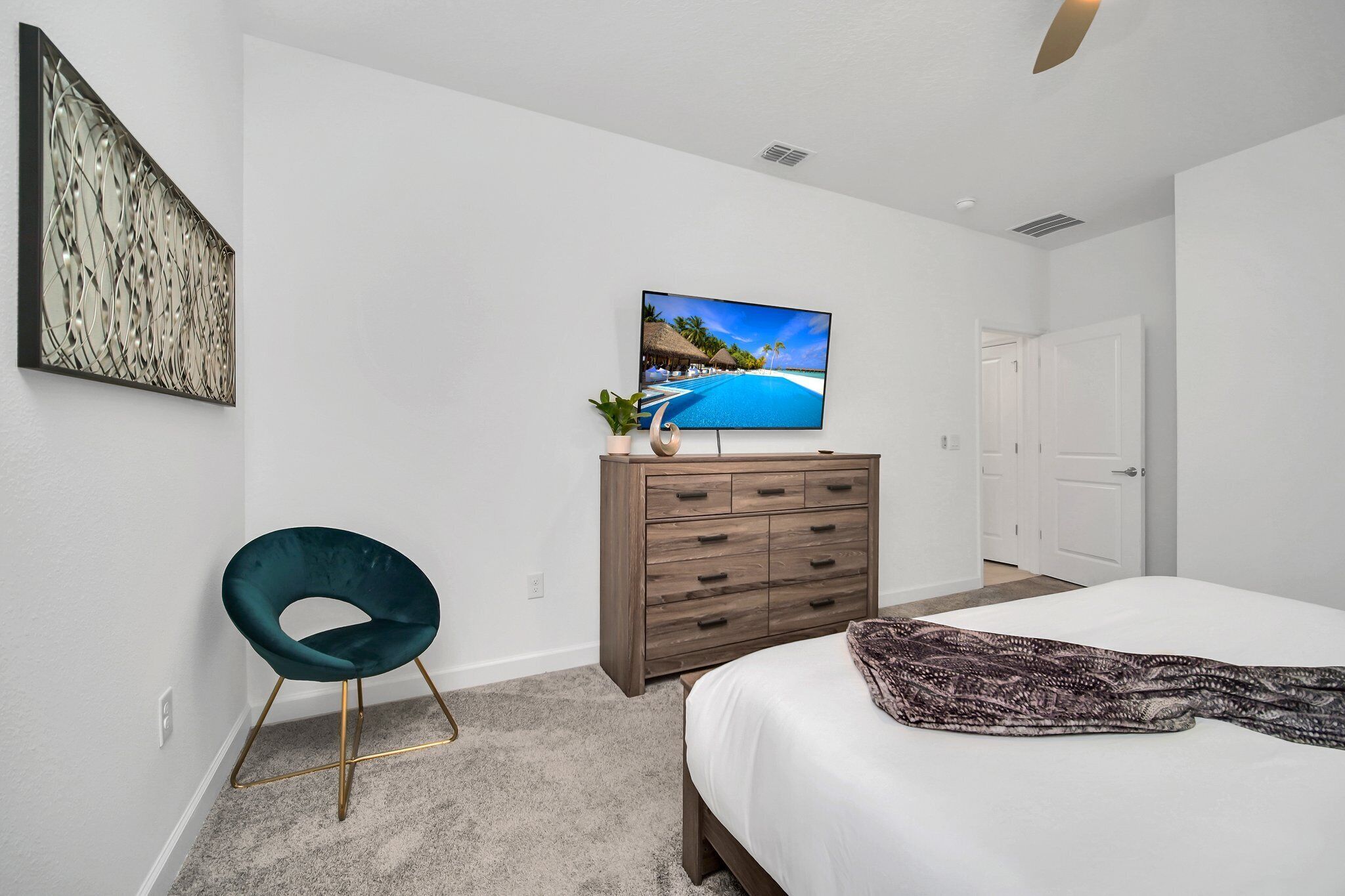 A bedroom with a bed, a TV on the wall, a wooden dresser, a green chair, and a painting on the wall.