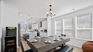A modern kitchen with white cabinets, a wooden dining table set with plates and glasses, a chandelier, and a large window with blinds. - Lakefront House with Ping Pong, Pool Table, & Front Patio Stunning Views (Nevada)