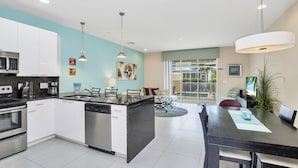 Une cuisine moderne avec des meubles blancs, un îlot central, des appareils en acier inoxydable, un éclairage suspendu et un coin repas avec une grande table et des chaises.