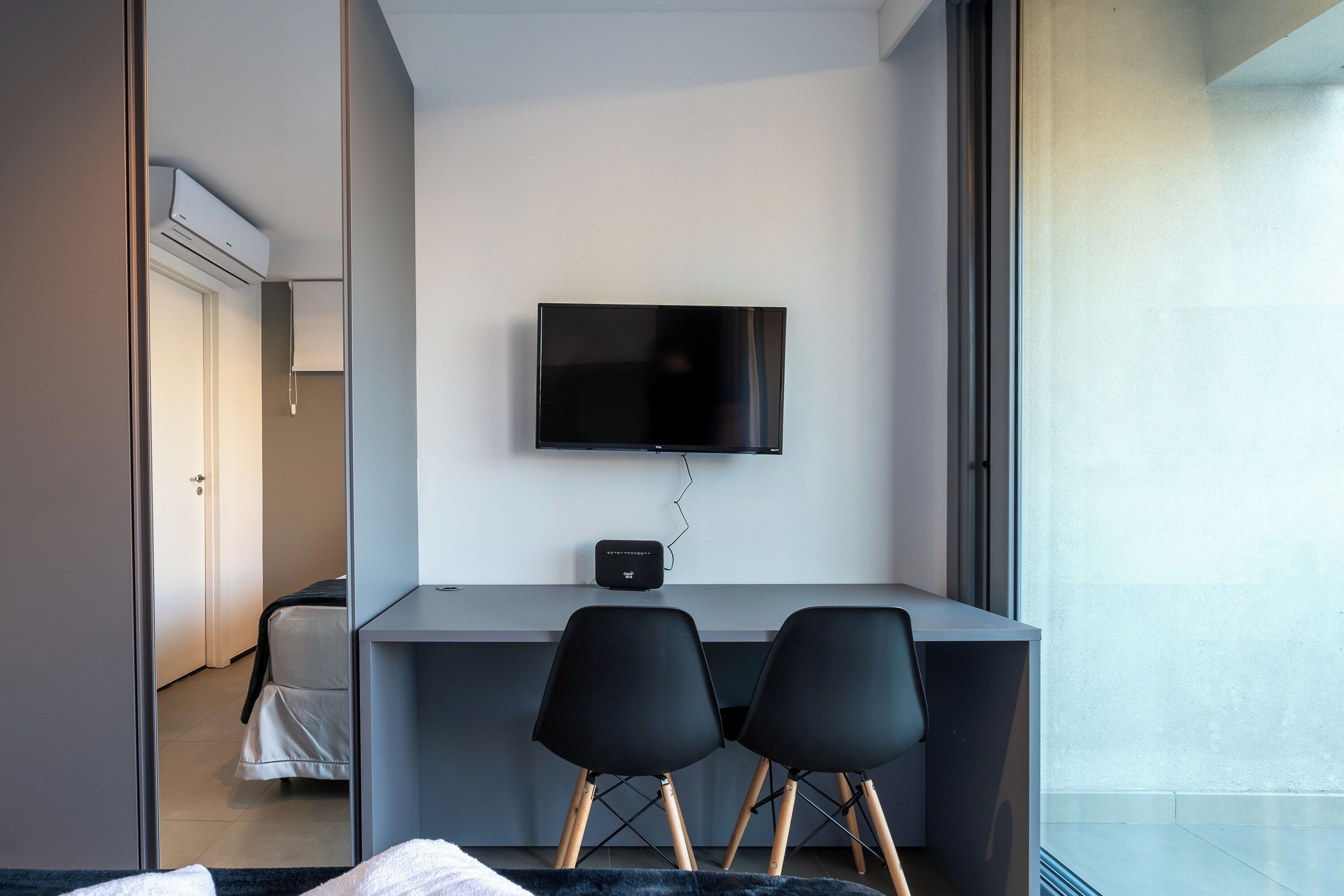 Une chambre d’hôtel moderne équipée d’un téléviseur à écran plat fixé au mur, de deux chaises noires à pieds en bois, d’un petit bureau et d’un lit recouvert d’une couverture de couleur sombre.