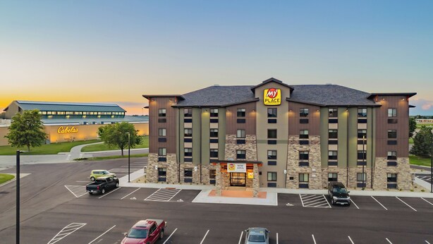 Un hotel de varios pisos con un letrero 'MY PLACE', un estacionamiento con varios coches y un estadio de béisbol al fondo.