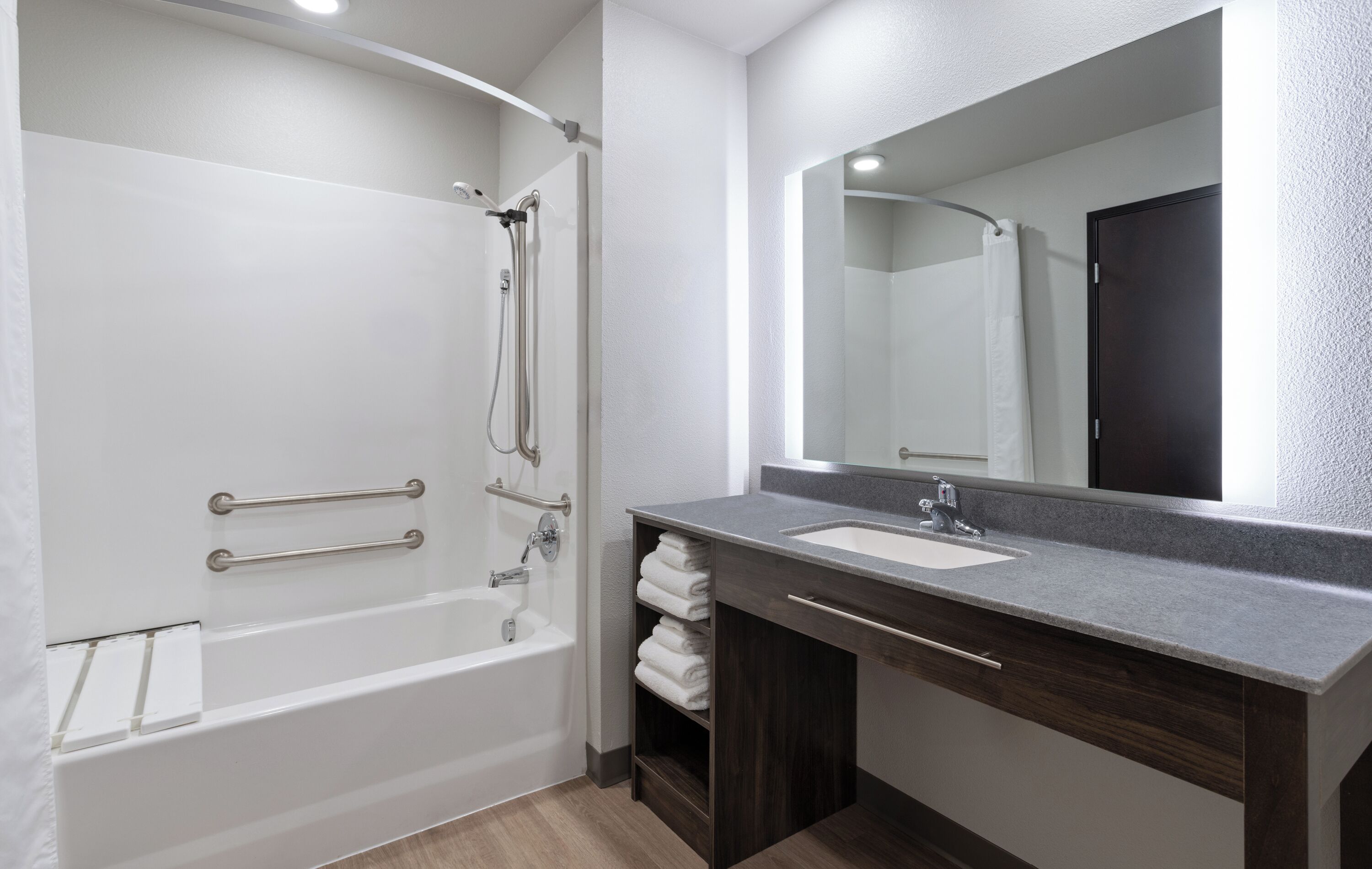 A modern bathroom with a walk-in shower, grab bars, a large mirror, and a dark wood vanity with a sink.