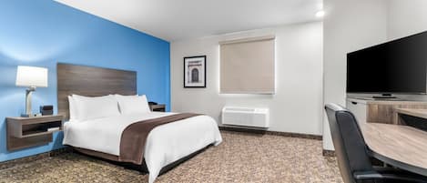 Quarto de hotel com uma cama, uma escrivaninha, uma cadeira, televisão, abajur e um quadro na parede.