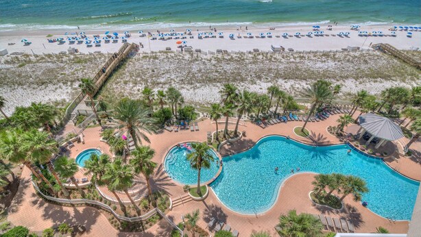 Un resort con una piscina grande, palmeras y una zona de playa con sombrillas.
