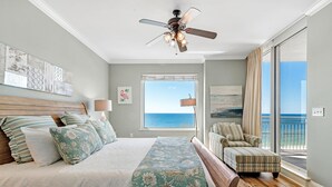 Un dormitorio con una cama grande, un ventilador de techo, una ventana con vista al océano y un balcón con una silla.