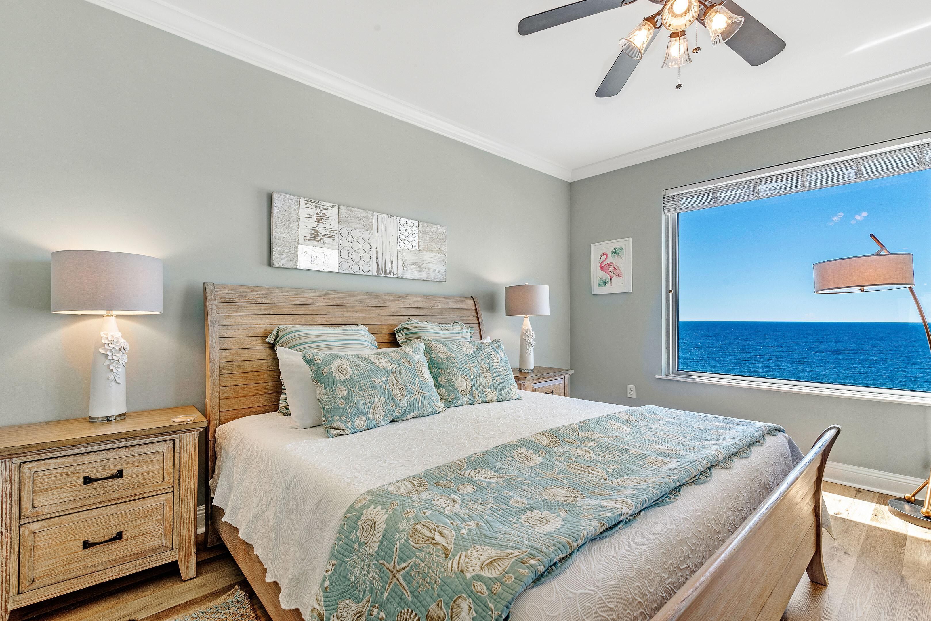 Quarto com cama, mesas de cabeceira, ventilador de teto e janela com vista para o oceano.