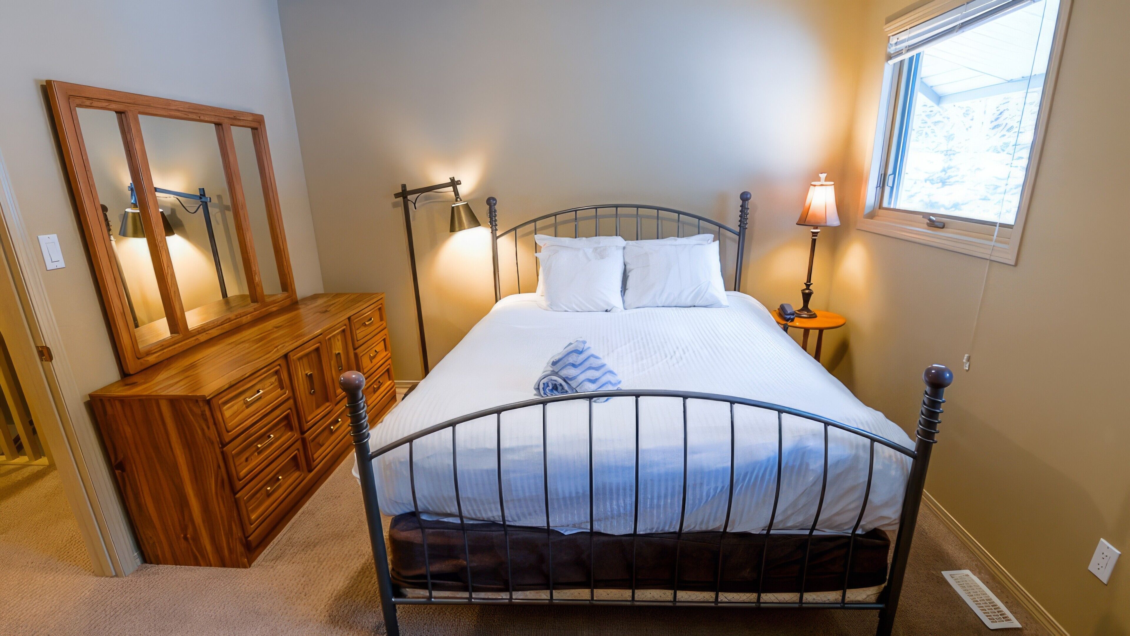 Een slaapkamer met een metalen bedframe, een houten dressoir, een spiegel, bedlampjes en een raam.