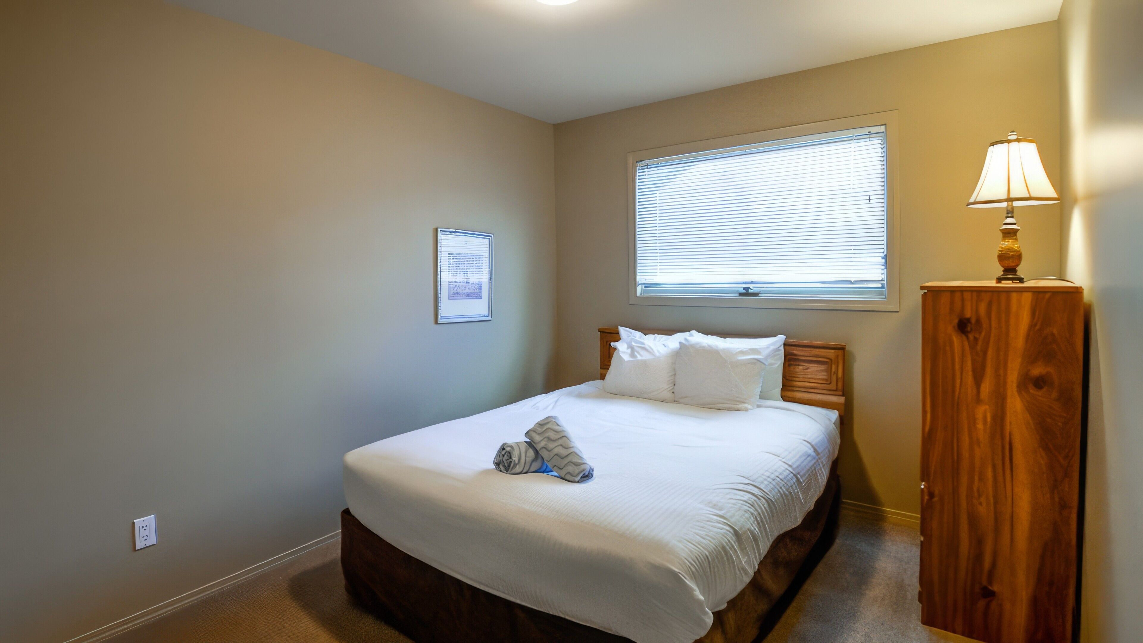 A bedroom with a single bed, a nightstand, a lamp, a window with blinds, and a framed document on the wall.