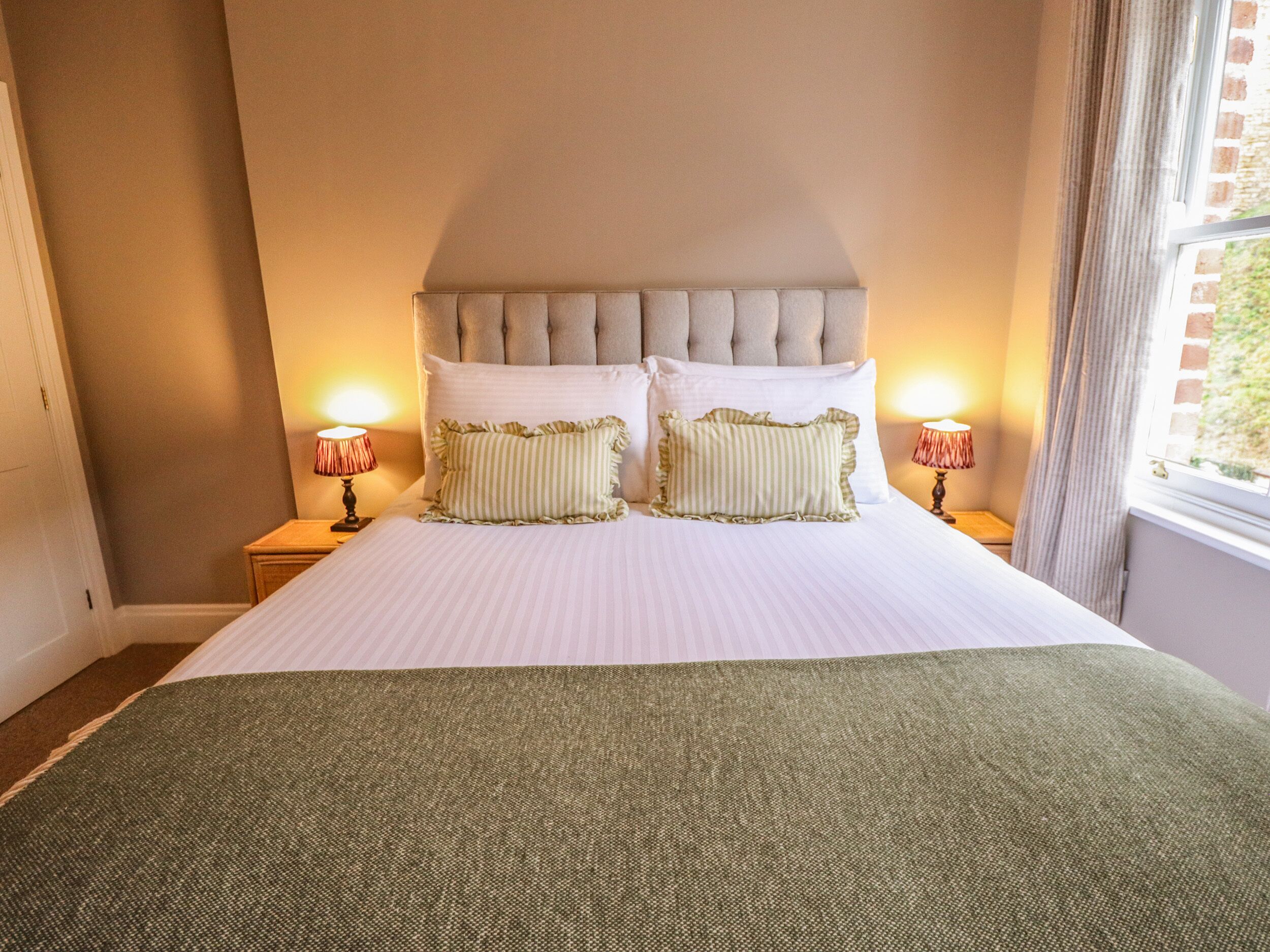 A neatly made bed with a tufted headboard, two bedside lamps, and a view of a brick wall and window.