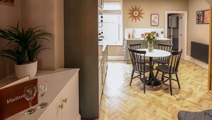 A dining area with a round table, chairs, and a potted plant. There is a kitchen in the background with a window and a clock on the wall. - 17 Union Road House (Lincoln)