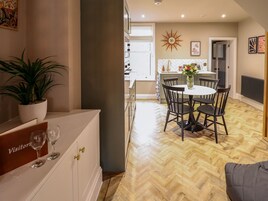Un comedor con una mesa redonda, sillas y una planta en maceta. Se observa una cocina al fondo con una ventana y un reloj en la pared.