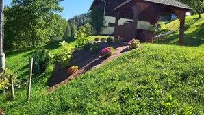 Una cabaña de madera con techo inclinado, rodeada de un exuberante jardín con diversas flores y una colina cubierta de césped.