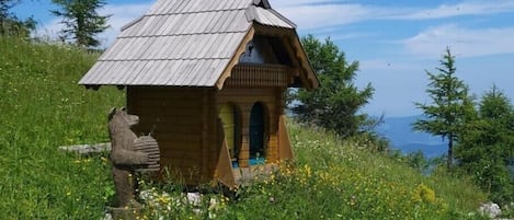 A wooden cabin with a sloped roof, situated on a grassy hill with a clear view of the sky and distant mountains.