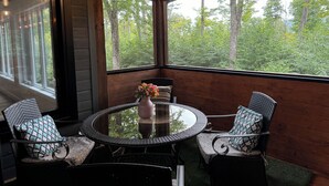 Una habitación con paneles de madera, una mesa redonda y cuatro sillas, ventanas amplias que dan a una vista del bosque, y una araña.