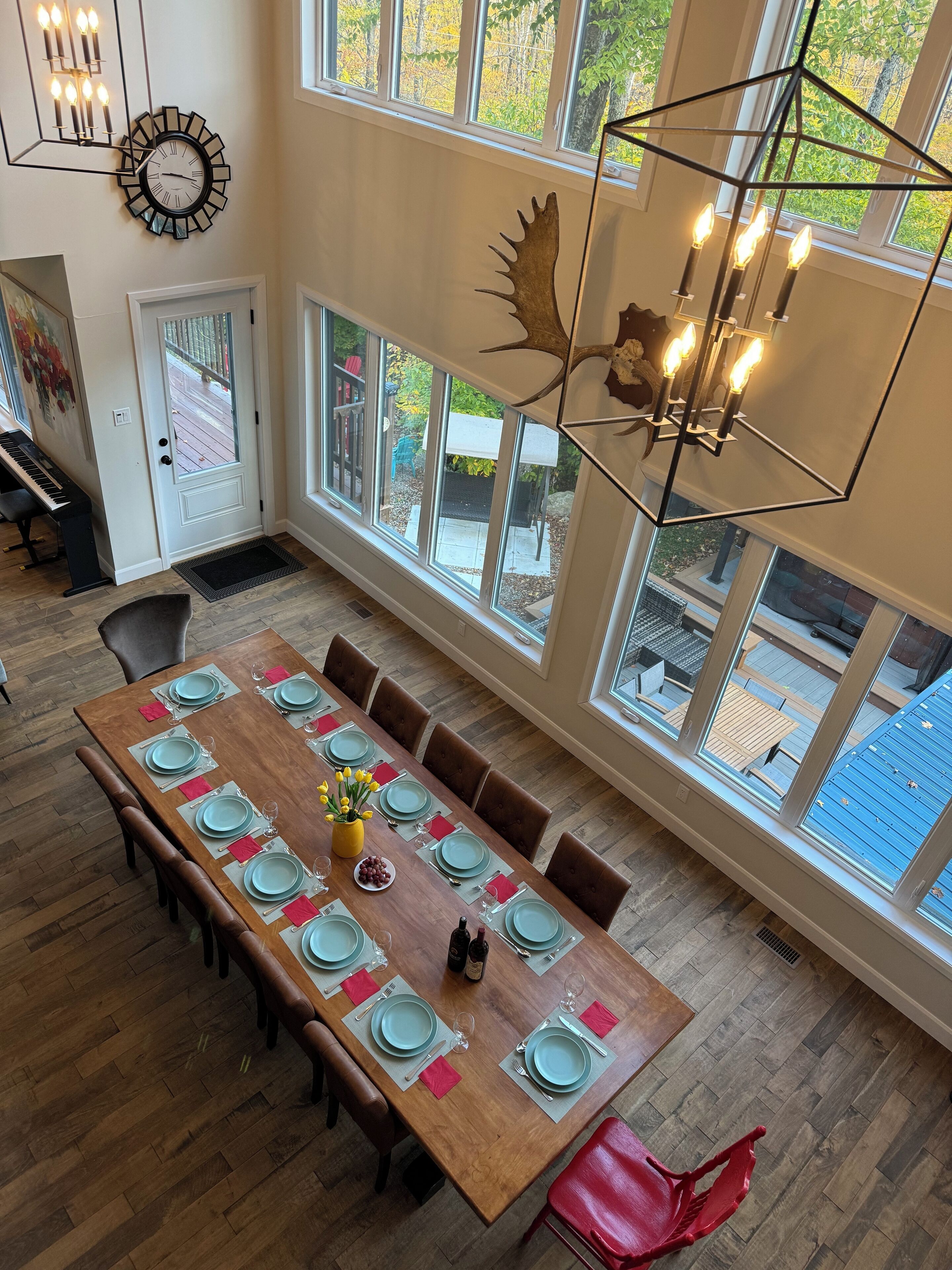 Uma sala de jantar com uma grande mesa de madeira posta para seis pessoas, cercada por seis cadeiras. Há um piano à esquerda e uma grande janela com vista para o exterior.