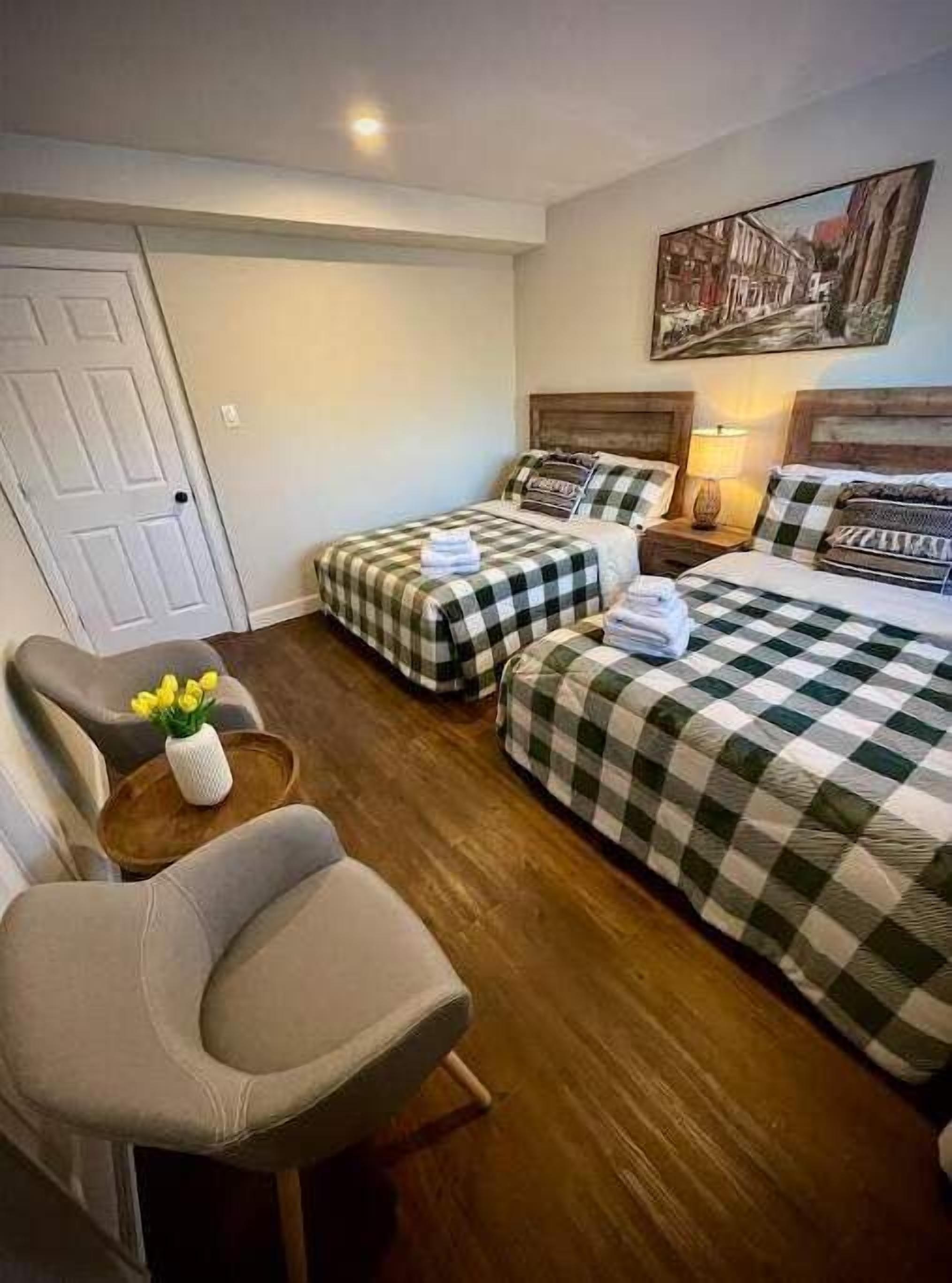 Une chambre d’hôtel avec deux lits, une chaise, une petite table sur laquelle est posé un vase de fleurs, et un tableau au mur.
