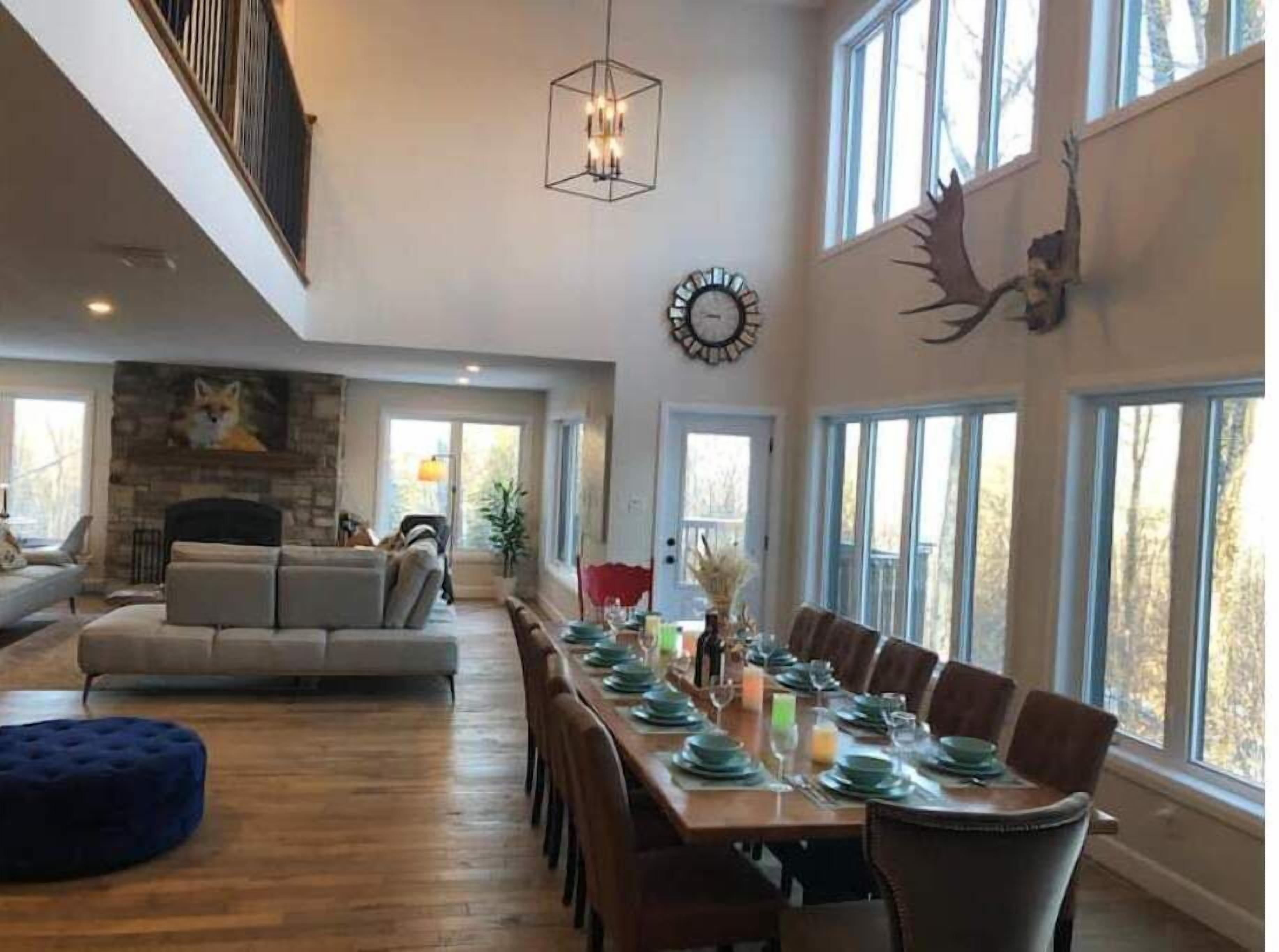 A spacious living room with a fireplace, a dining area with a long table set for six, and large windows offering a view of the outdoors.