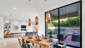 A modern dining area with a wooden table set for four, surrounded by black chairs. A large window offers a view of an outdoor patio with seating. - LA Villa by Avantstay Walk to The Grove LA (Los Angeles)